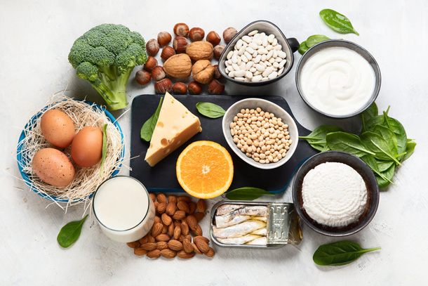 Es liegen unterschiedliche Lebensmittel auf einem weissen Tisch, die viel Calcium enthalten. Eier, Broccoli, Nüsse, Bohnen, Hülsenfrüchte, Frischkäse, Hartkäse & Sardinen in einer Dose.