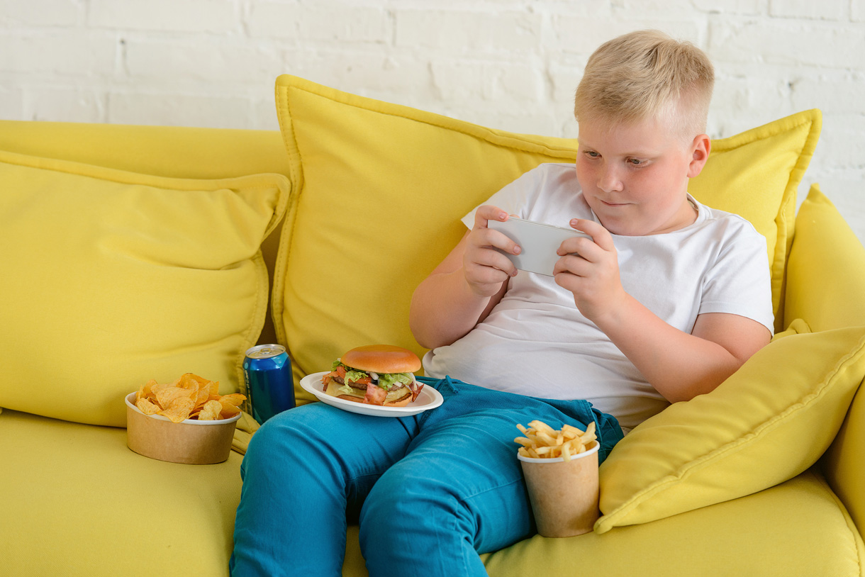 Gesunde Ernährung bei Kindern stellt die Weichen fürs ganze Leben. (Foto: 123RF, yuragolub) Ein übergewichtiger Junge sitzt mit dem Handy auf dem Sofa. Um ihn herum steht ein Süssgetränk und fast Food.