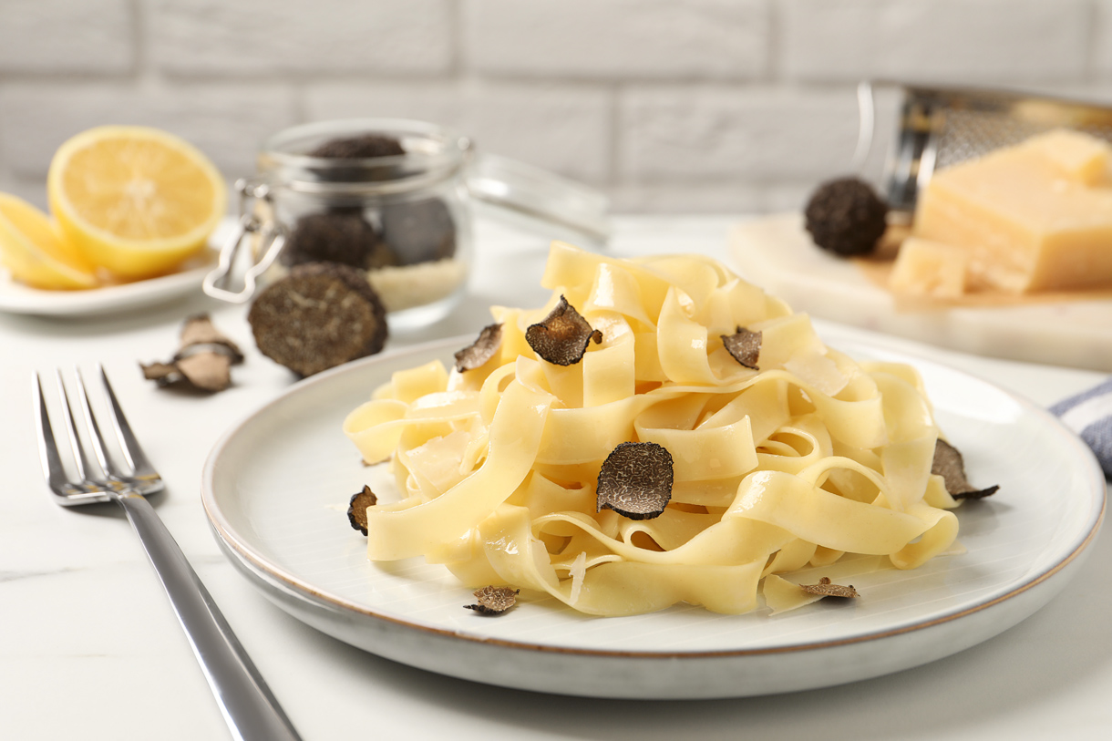 Trüffelpasta (Foto: 123RF, liudmilachernetska) Ein fertiger Teller frischer Pasta stehen auf einem Tisch. Über den Nudeln ist frischer Trüffel gehobelt worden. Man sieht den Trüffel noch neben dem Teller liegen. Im Hintergrund sieht man noch eine aufgeschnittene Zitrone und Parmesankäse.