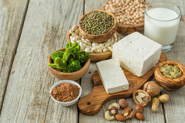 Auf einem Holztisch liegen in diversen Schüsselchen Linsen, Bohnen, Nüsse und Samen. Daneben steht ein Glas Pflanzenmilch und ein Tofu-Block liegt im Zentrum auf einem Holzbrett.