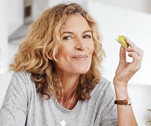 Einfache Diät-Tipps für die Wechseljahre (Foto: 123RF, peopleimages12) Eine blonde Frau mit lockigem Haar stützt sich in Ihrer Küche gemütlich auf die Kücheninsel und ist einen frisch geschnitten Apfelschnitz.
