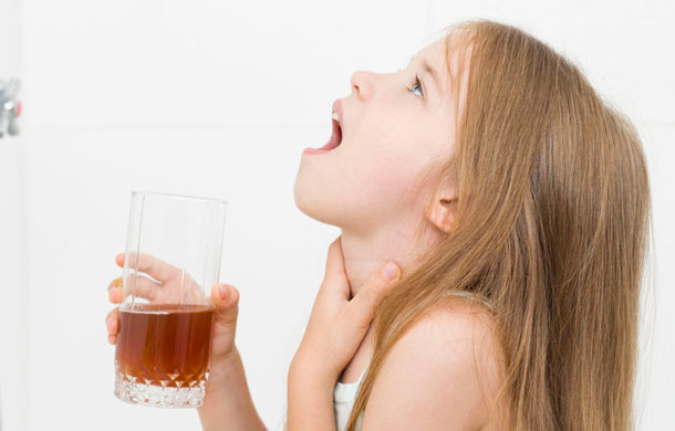 Ein Mädchen ist gerade dabei zu gurgeln. Der Kopf ist nach hinten geneigt und der Mund steht weit offen. In der rechten Hand hält sie ein Glas mit einer braunen Flüssigkeit. (Gurgel-Lösung)