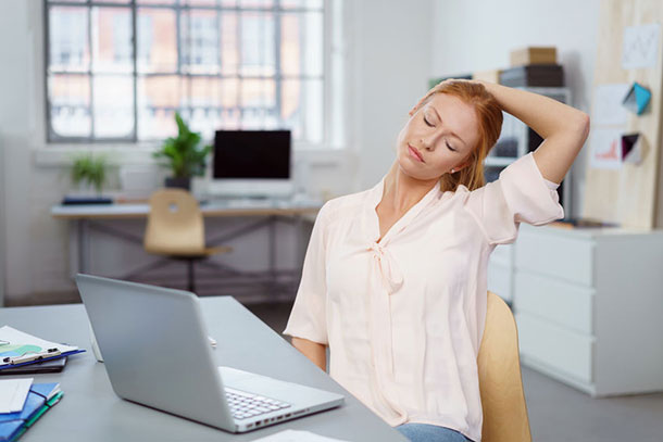 Une jeune femme est assise devant son ordinateur portable dans le bureau, essayant de détendre son cou. Elle tend la tête vers la gauche et la tient avec son bras gauche.