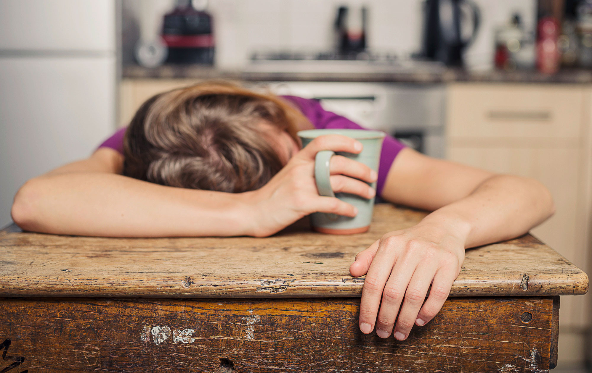Müdigkeit ist ein häufiger Konsultationsgrund in ambulanten Praxien und heute wohl in der Schweiz die häufigste Indikation zur Eisentherapie. (Foto: iStock, lolostock) Eine Frau sitzt am Küchentisch, ihr Oberkörper liegt vor erschöpfung flach auf dem Tisch. Mit einer Hand hält sie eine Tasse fest.