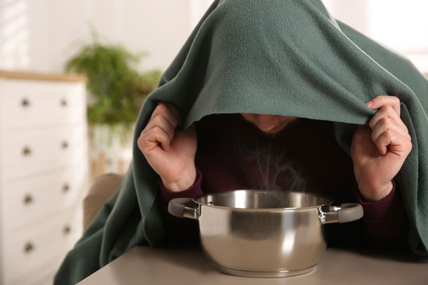Eine Person sitzt über einem dampfenden Topf, an einem Tisch, und inhaliert die Dämpfe mit einem Tuch über dem Kopf bewusst ein. 