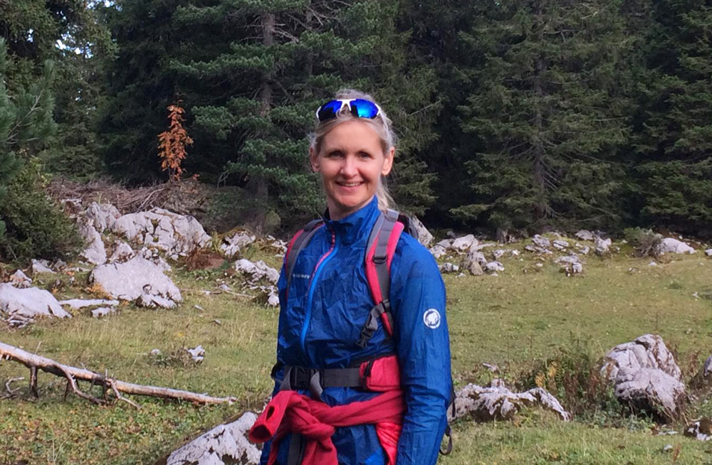 Sonja Schröder unterwegs auf Wanderschaft. Da Bild zeigt Sonja Schröder inmitten von grüner Natur. Sie steht in Wanderkleidung auf einer Alp-Wiese. Im Hintergrund ist ein Wald.